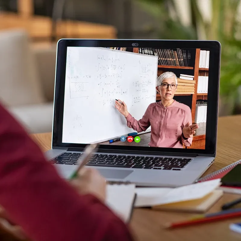 A woman sitting a desk taking notes while watching a lecture on a laptop